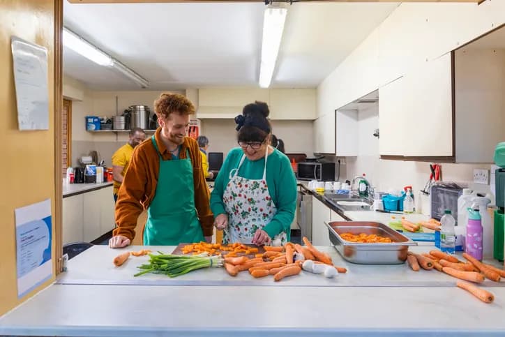 A man and a woman in aprons stand at a counter in a kitchen, chopping carrots and scallions, while other people work in the background.