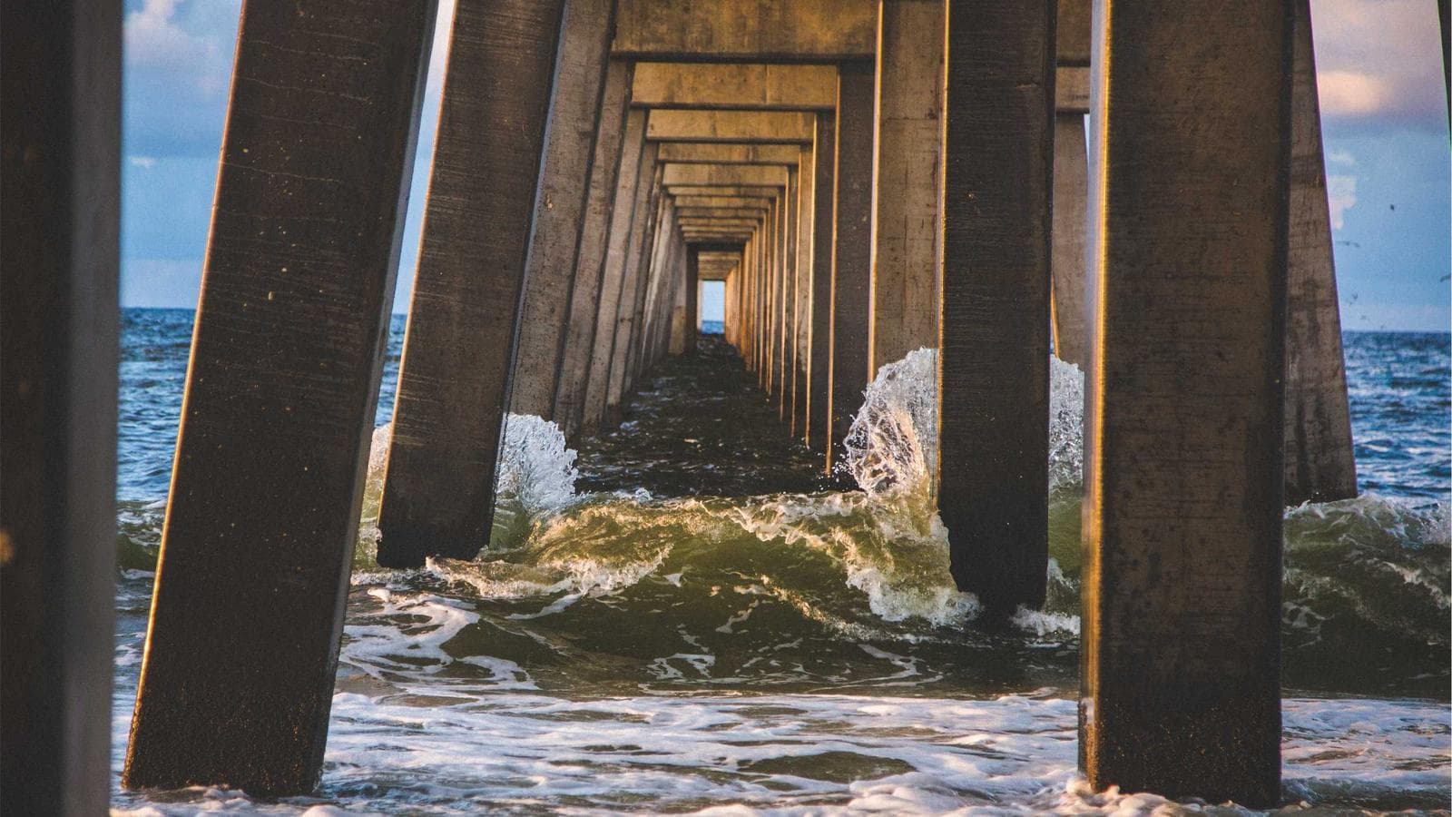 Waves crash against a concrete pier creating an infinite tunnel effect that narrows to a bright opening.